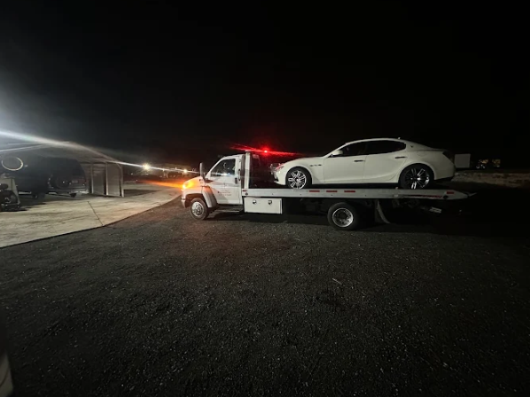 Late-night vehicle recovery of a white sedan on a flatbed tow truck